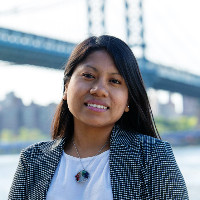 photo of Deysi Flores, looking at the camera with long brown hair and medium brown skin, in a white blouse and black patterned blazer, with the Brooklyn Bridge in the background